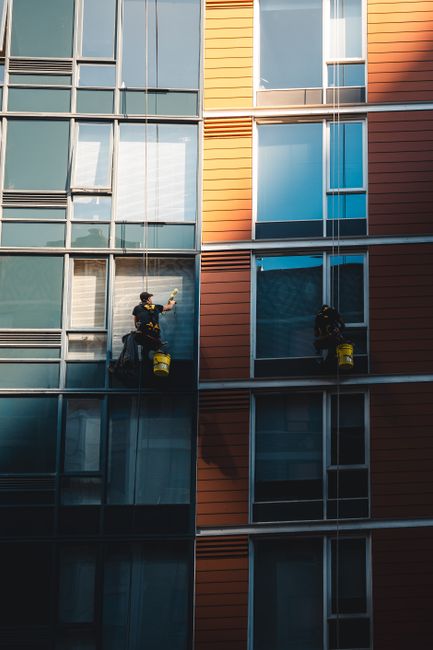 Two window cleaners are suspended mid-air on the exterior of a modern high-rise building, cleaning large glass panels. They are wearing safety harnesses and work gear, including helmets and harnesses 