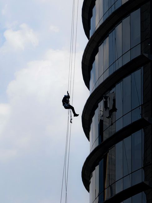 Two window cleaners are suspended mid-air on the exterior of a modern high-rise building, cleaning large glass panels. They are wearing safety harnesses and work gear, including helmets and harnesses 