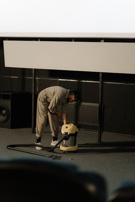 A person vacuuming an ornate, patterned area rug in a residential living room, using a yellow and black vacuum cleaner. The rug features a floral design with beige, green, and orange tones. The indivi