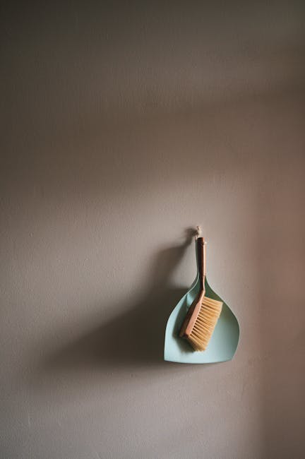 A dustpan and brush hanging on a plain white wall. The dustpan is made of light blue plastic and is positioned vertically, with the handle of the brush resting inside it. The brush has light-colored b