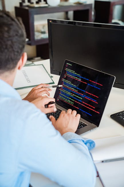 A person seated at a desk working on a laptop displaying colorful lines of code, with a large computer monitor, a closed notepad, and a black wireless keyboard visible on the white desk. The room appe